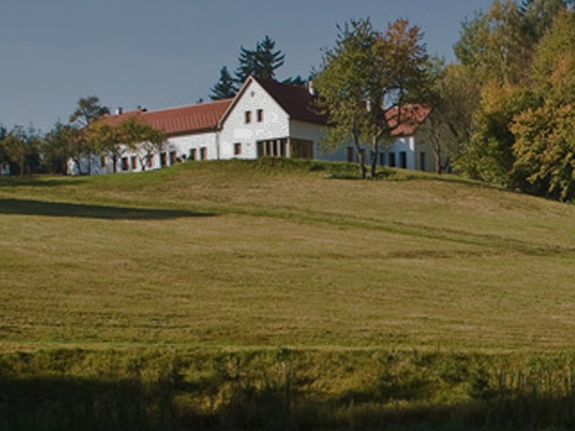 Das weiße Hofgebäude des Rosenhofs mit rotem Ziegeldach steht auf einer Kuppe über einer weiten Wiese, dahinter Wald.
